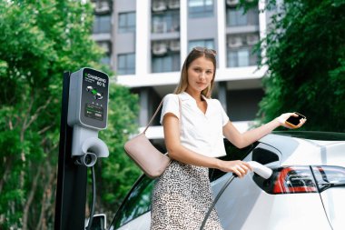 Genç bir kadın yazın yeşil sürdürülebilir şehir bahçesinde EV elektrikli arabasıyla seyahat ediyor. Kentsel sürdürülebilirlik yaşam tarzı yeşil temiz yeniden şarj edilebilir enerji elektrikli BEV araç iç organları