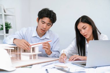 Profesyonel Asyalı erkek mimar ev modelinin uzunluğunu ölçmek için cetvel kullanırken, beyaz tenli genç meslektaşı ev modeliyle toplantı masasındaki verileri analiz etmek için dizüstü bilgisayar kullanıyor. Tertemiz..