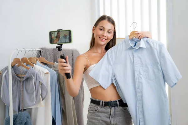 Young woman making fashion video content for social media. Blogger smiles to camera using selfie stick and light ring while making persuasive online clothing sell to audience or followers. Unveiling
