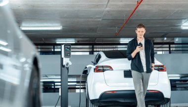 Genç adam EV elektrikli arabasıyla alışveriş merkezinin otoparkına gidiyor şehir merkezindeki şarj oluyor yeşil, temiz, yeniden şarj edilebilir elektrikli araç iç organları ile kentsel sürdürülebilirlik yaşam tarzını gösteriyor