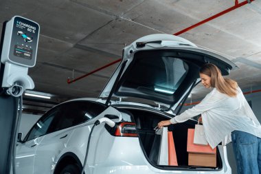 Genç bir kadın EV elektrikli arabasıyla alışveriş merkezinin otoparkına gidiyor şehir merkezinde elektrik iç organlarının yeşil temiz şarj edilebilir enerji ile sürdürülebilirlik yaşam tarzını gösteriyor.