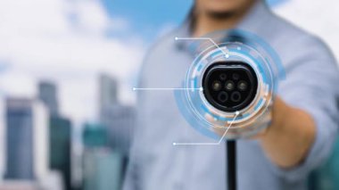 Kameranın önüne odaklanmış EV şarj cihazı bulanık şehir manzarası ve hızlandırılmış bulut arkaplanlı dijital akıllı batarya durum hologramını gösterir. Alternatif enerji kullanan EV araba şarjı. İnceleyin