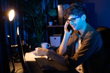 Genç iş adamı akıllı telefondan müşteriyi ya da iş arkadaşını arıyor, neon modern ofisteki yaratıcı sunum surround çalışma masasının bilgisayar ekranına bakıyor. Yeni proje kavramı. Toplayıcı.