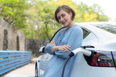 Yeşil kırsal kesimde tatil gezisi. Güzel genç bir kadın alternatif enerjiyle elektrikli aracı şarj ediyor. Çevre dostu seyahat akıllı EV araba. Kalıcı