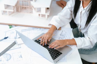 Profesyonel Asyalı erkek mimar ev modelinin uzunluğunu ölçmek için cetvel kullanırken, beyaz tenli genç meslektaşı ev modeliyle toplantı masasındaki verileri analiz etmek için dizüstü bilgisayar kullanıyor. Tertemiz..