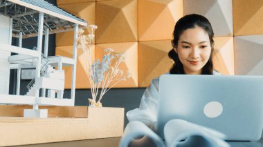 Genç ve güzel mimar mühendisin portresi çatı malzemelerini kontrol ediyor ve ofisteki masaya yerleştirilmiş proje planı ve ev modeliyle dizüstü bilgisayar kullanıyor. Yaratıcı iş tasarımı konsepti. Yönlendirici.