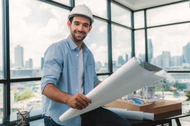 Ev modelinde proje planını ve gökdelenli mimar ekipmanlarını okurken kameraya bakan profesyonel mimar mühendisinin portresi. Mimarlık stüdyosuyla çevrili. İzleme.