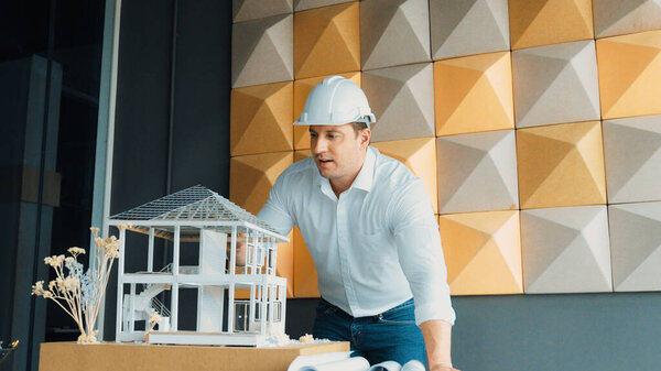 Closeup portrait of caucasian male architect engineer inspect and check house model construction with project plan and architectural model placed on table. Business design concept. Manipulator.