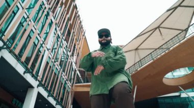 Hip hop tarzında düşük açılı hipster pozu alışveriş merkezi veya bina manzarası ile çevrilidir. Kameraya bakarken Asyalı hip hop koreografı break dans yapıyor. Açık hava sporu 2024 hiphop..