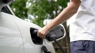 Adam EV şarj aleti takıyor ve yeşil park ve yeşillik arka planında akıllı şarj istasyonu olan elektrikli arabayı şarj ediyor. Çevre koruma için temiz enerjiden güç alan çevre dostu EV araba. İnceleyin