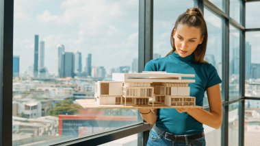 Genç akıllı mimar mühendisi ev modelini incelerken mimari modeli elinde tutuyor. Şehir manzaralı pencerenin yanında dururken profesyonel iç mimar ev inşaatını kontrol ediyor. İzleme