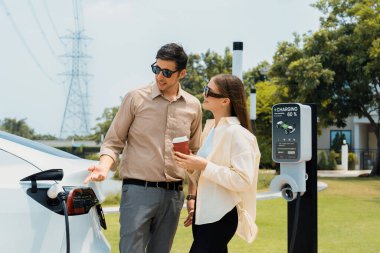 Elektrik faturası için kahve ödeyen genç bir çift elektrik santralindeki EV araba aküsünü şarj ederken elektrik şebekesine bağlı elektrik endüstrisi de çevre dostu otomobil seyahati için kullanılıyor. Yararlı