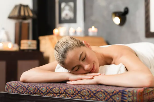 Beautiful relaxing caucasian woman lie on spa bed in front of wooden sauna cabinet. Young and healthy woman waiting for massage treatment in warm traditional spa room. Tranquility.