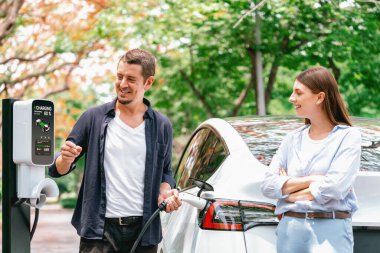 Sonbahar gezisi sırasında sonbahar ormanı EV arabasında elektrikli araba için şarj olan sevimli genç çift. Sonbahar tatilinde çevre dostu seyahat. Yükselt