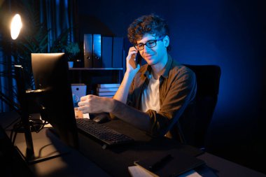 Genç iş adamı akıllı telefondan müşteriyi ya da iş arkadaşını arıyor, neon modern ofisteki yaratıcı sunum surround çalışma masasının bilgisayar ekranına bakıyor. Yeni proje kavramı. Toplayıcı.