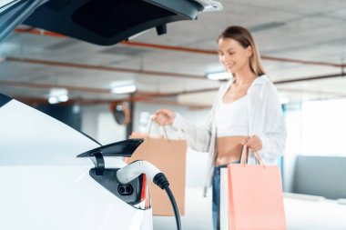 Genç bir kadın EV elektrikli arabasıyla alışveriş merkezinin otoparkına gidiyor şehir merkezinde elektrik iç organlarının yeşil temiz şarj edilebilir enerji ile sürdürülebilirlik yaşam tarzını gösteriyor.