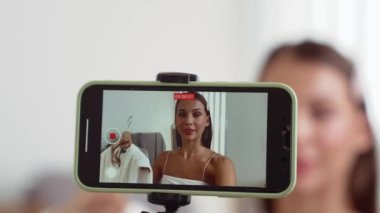 Genç sosyal medyum kadın moda videosu yapıyor. Blogcu kameraya selfie çubuğu ve ışık halkası kullanarak gülümserken, aynı zamanda ikna edici çevrimiçi kıyafetlerin izleyicilere veya takipçilere vlog satmasını sağlıyor. Düzenle