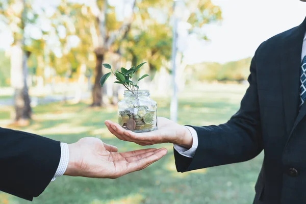 Concept of sustainable money growth investment with glass jar filled with money savings coins with businesspeople as eco-friendly financial investment nurtured with nature and healthy retirement. Gyre