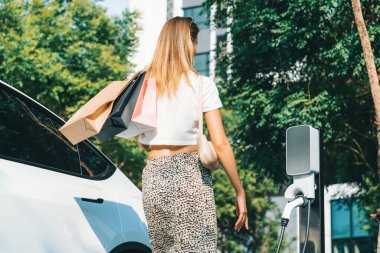 Genç bir kadın yazın yeşil sürdürülebilir şehir bahçesinde EV elektrikli arabasıyla seyahat ediyor. Kentsel sürdürülebilirlik yaşam tarzı yeşil temiz yeniden şarj edilebilir enerji elektrikli BEV araç iç organları