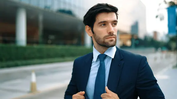 Portrait of business man adjust tie while walking along the road. Manager wearing suit while looking around the city. Investor check himself before going to interview. Blurred background. Exultant.