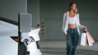 Genç bir kadın EV elektrikli arabasıyla alışveriş merkezinin otoparkına gidiyor şehir merkezinde elektrik iç organlarının yeşil temiz şarj edilebilir enerji ile sürdürülebilirlik yaşam tarzını gösteriyor.