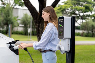 Açık yeşil şehir parkı manzaralı EV şarj istasyonundan genç kadın EV elektrikli araçları şarj ediyor. Çevre dostu şehir ulaşımı ve çevre dostu EV araba seyahati. Yükselt