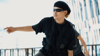 Arkadaşı gökyüzü dövüşçüsünün arkasında dans ederken Asyalı hipster kameraya bakıyor. Bir grup sokak dansçısı kameraya bakarken break dans yapıyor. Açık hava sporu 2024. ÇalışmaComment.