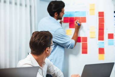 Çeşitli ofis çalışanlarından oluşan bir grup, toplantı odasındaki beyaz tahtaya yapışkan bir notla stratejik iş pazarlama planlaması üzerine beyin fırtınası yapıyor. İş yerinde verimli bir takım çalışması. Tedbirli