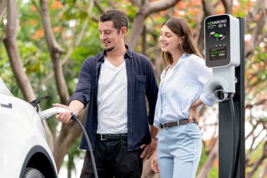 Sonbahar gezisi sırasında sonbahar ormanı EV arabasında elektrikli araba için şarj olan sevimli genç çift. Sonbahar tatilinde çevre dostu seyahat. Yükselt