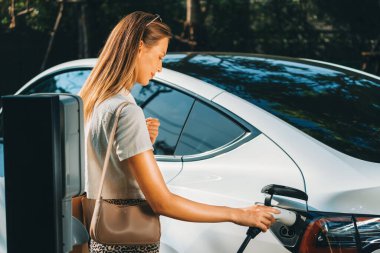 Genç bir kadın yazın yeşil sürdürülebilir şehir bahçesinde EV elektrikli arabasıyla seyahat ediyor. Kentsel sürdürülebilirlik yaşam tarzı yeşil temiz yeniden şarj edilebilir enerji elektrikli BEV araç iç organları