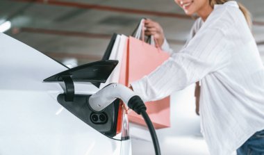 Genç bir kadın EV elektrikli arabasıyla alışveriş merkezinin otoparkına gidiyor şehir merkezinde elektrik iç organlarının yeşil temiz şarj edilebilir enerji ile sürdürülebilirlik yaşam tarzını gösteriyor.