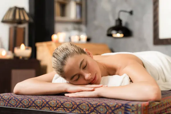 Beautiful relaxing caucasian woman lie on spa bed in front of wooden sauna cabinet. Young and healthy woman waiting for massage treatment in warm traditional spa room. Tranquility.