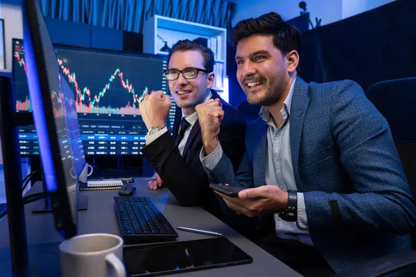Two stock exchange traders comparing dynamic investment graph via phone and pc with high profit in currency rate. Showing financial benefit data on monitor screen in neon light at workplace. Sellable.