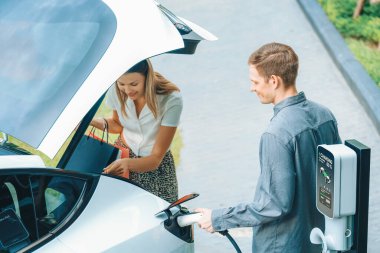 Genç çift yazın yeşil sürdürülebilir şehir bahçesinde EV elektrikli araba ile seyahat ederken, yeşil temiz yeniden şarj edilebilir elektrikli araç iç organları ile kentsel sürdürülebilirlik yaşam tarzını gösteriyor.