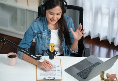 Sosyal medyada kulaklıkla canlı yayın yapan Asyalı sunucu modern stüdyoda yaşam koçluğu veya pazarlama dinleyicilerini selamlıyor. Taktik.