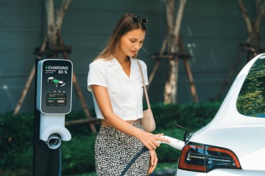 Genç bir kadın yazın yeşil sürdürülebilir şehir bahçesinde EV elektrikli arabasıyla seyahat ediyor. Kentsel sürdürülebilirlik yaşam tarzı yeşil temiz yeniden şarj edilebilir enerji elektrikli BEV araç iç organları