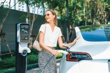 Genç bir kadın yazın yeşil sürdürülebilir şehir bahçesinde EV elektrikli arabasıyla seyahat ediyor. Kentsel sürdürülebilirlik yaşam tarzı yeşil temiz yeniden şarj edilebilir enerji elektrikli BEV araç iç organları