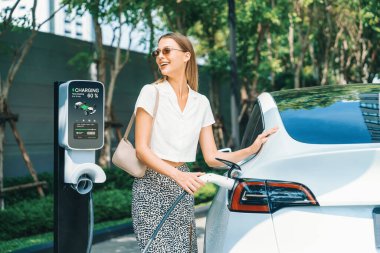 Young woman travel with EV electric car charging in green sustainable city outdoor garden in summer. Urban sustainability lifestyle by green clean rechargeable energy of electric BEV vehicle innards clipart