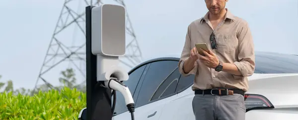 Man pay for electricity with smartphone while recharge EV car battery at charging station connected to electrical lower grid tower for eco friendly car utilization. Panorama Expedient