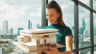 Genç akıllı mimar mühendisi ev modelini incelerken mimari modeli elinde tutuyor. Şehir manzaralı pencerenin yanında dururken profesyonel iç mimar ev inşaatını kontrol ediyor. İzleme