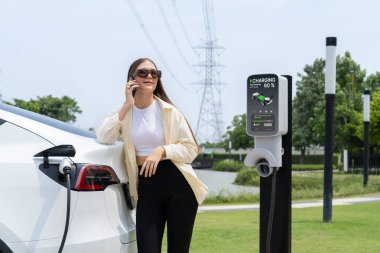 EV araba aküsünü şarj eden genç bir kadın elektrik şebekesine bağlı elektrik santralinde çevre dostu araç kullanımı için elektrik endüstrisi olarak telefonla konuşurken. Yararlı