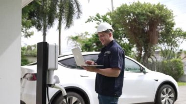 Evde EV şarj istasyonu kurulumu üzerinde çalışan nitelikli teknisyen, EV için dizüstü bilgisayarla şarj sisteminde sorun çıkarma ve yapılandırma ayarları yapıyor. Titiz