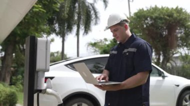 Evde EV şarj istasyonu kurulumu üzerinde çalışan nitelikli teknisyen, EV için dizüstü bilgisayarla şarj sisteminde sorun çıkarma ve yapılandırma ayarları yapıyor. Titiz