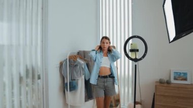 Genç sosyal medyum kadın moda videosu yapıyor. Blogcu kameraya selfie çubuğu ve ışık halkası kullanarak gülümserken, aynı zamanda ikna edici çevrimiçi kıyafetlerin izleyicilere veya takipçilere vlog satmasını sağlıyor. Düzenle