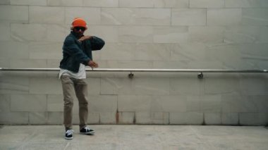 Asyalı akıllı hipster, kulaklık ve şık kıyafetler giyip hip hop şarkıları dinlerken gri arka planlı enerjik break dans gösterisi yapıyor. Açık hava sporu 2024. Hip hop hareketi. Yumuşak bir şekilde..