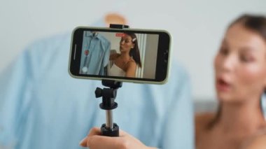 Genç sosyal medyum kadın moda videosu yapıyor. Blogcu kameraya selfie çubuğu ve ışık halkası kullanarak gülümserken, aynı zamanda ikna edici çevrimiçi kıyafetlerin izleyicilere veya takipçilere vlog satmasını sağlıyor. Düzenle
