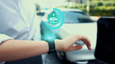 İş kadını, istasyonun otoparkından şarj olurken akıllı telefon hologramındaki EV aküsünün durumunu kontrol ediyor. EV teknolojisi adaptasyonu için temiz enerjinin gelecekteki yaşam tarzı. İnceleyin