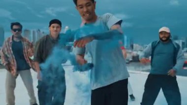 Bir grup çok kültürlü hippi çatıda işaret fişeği çalıyor. Zenci dumanı tutan çekici bir sokak dansçısı hip-hop müzik eşliğinde break dans yapıyor. Açık hava sporu 2024. Sokak dansı. hiphop.