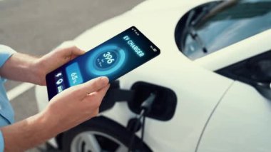 Akıllı telefon ekran bataryasını elinde tutan iş adamı akıllı EV mobil uygulama ile otomobil parkındaki şarj istasyonundan elektrik şarj eden çevre dostu EV otomobil. İnceleyin