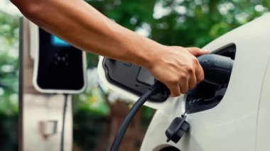 Adam EV şarj aleti takıyor ve yeşil park ve yeşillik arka planında akıllı şarj istasyonu olan elektrikli arabayı şarj ediyor. Çevre koruma için temiz enerjiden güç alan çevre dostu EV araba. İnceleyin